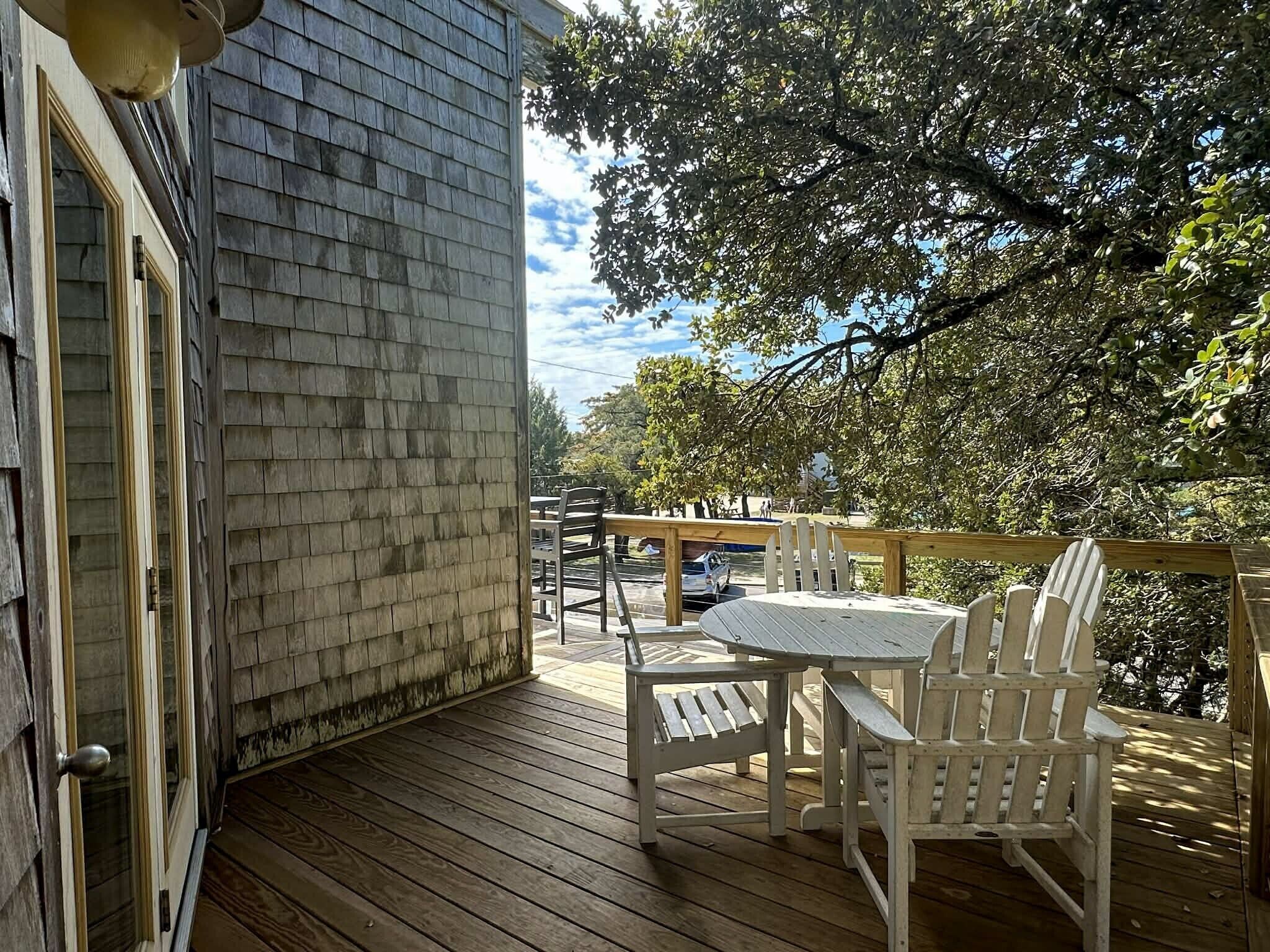 Une terrasse en bois avec une table et des chaises, une porte vitrée et une vue sur une rue bordée d’arbres.