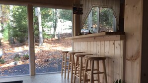 Un coin bar en bois avec trois tabourets, une plante en pot et une vue sur une zone boisée grâce à de grandes fenêtres.