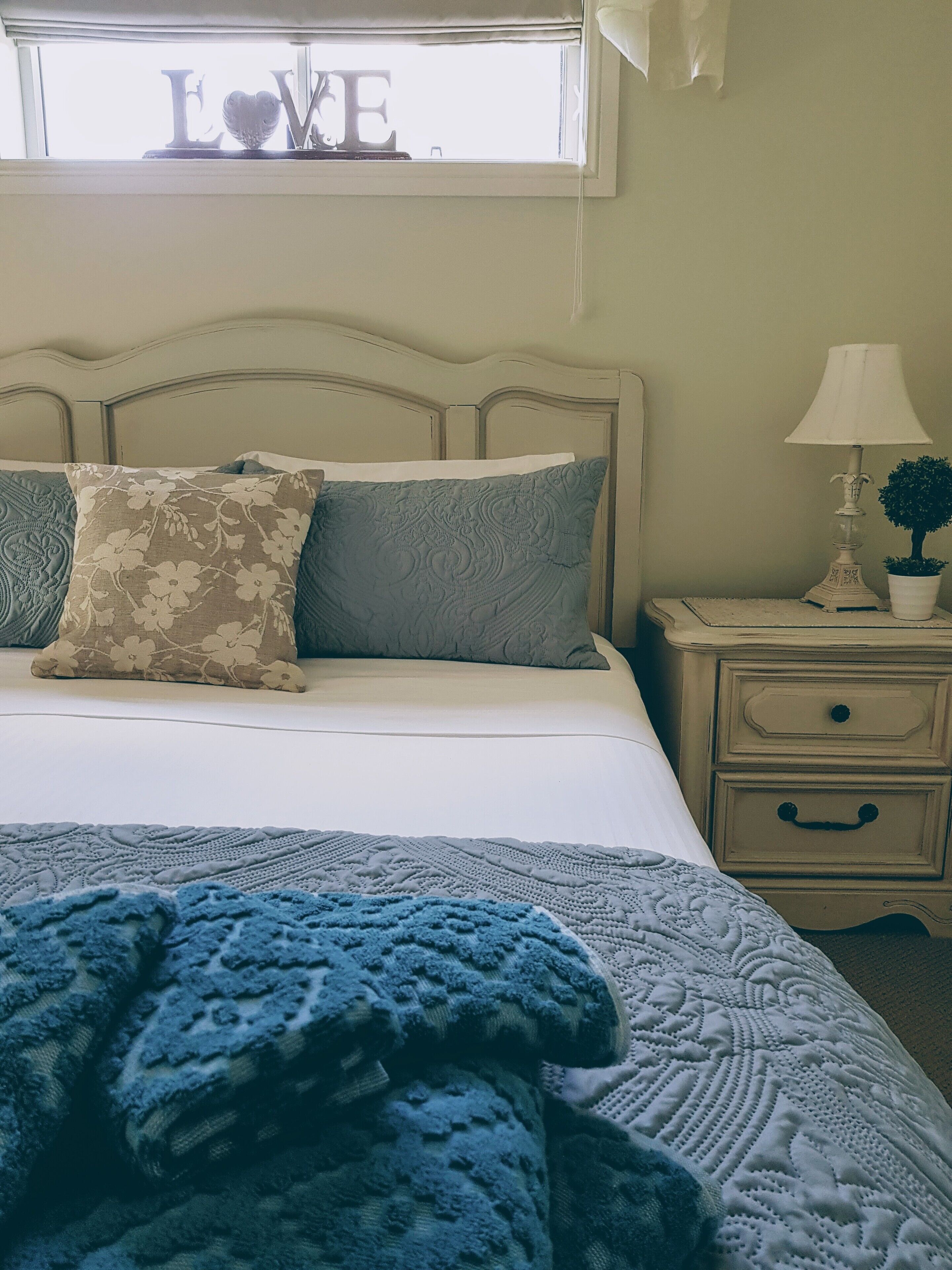 Ein ordentlich bezogenes Bett mit floralen und strukturierten Kissen, einer blauen Decke und einem Nachttisch mit Lampe und einer kleinen Pflanze.