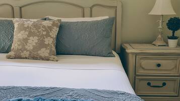 Ein ordentlich bezogenes Bett mit floralen und strukturierten Kissen, einer blauen Decke und einem Nachttisch mit Lampe und einer kleinen Pflanze.