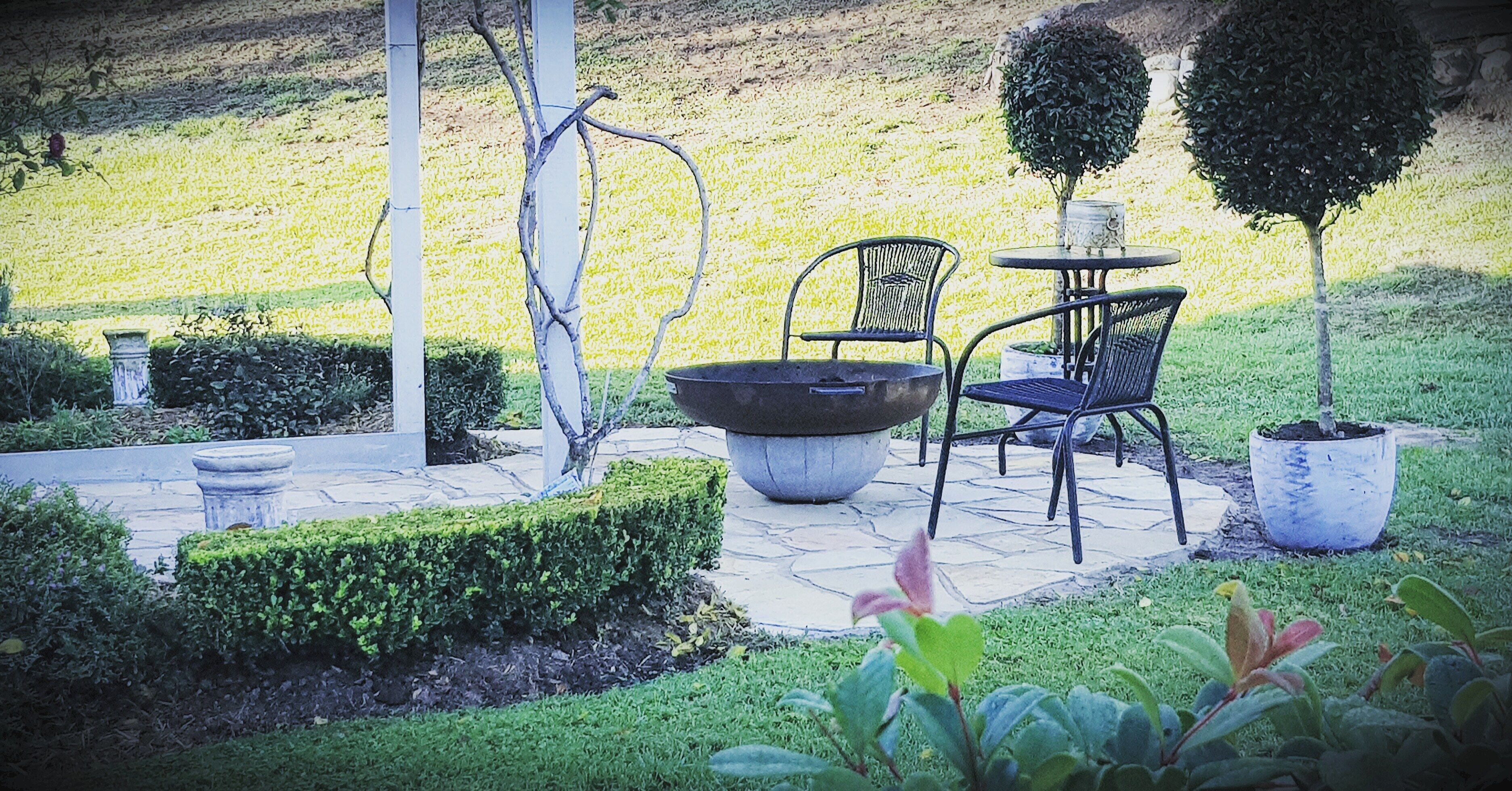 A patio with a table and chairs, surrounded by well-maintained hedges and a small tree.