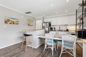 A modern kitchen with white cabinetry, a granite countertop, and a dining area with wicker chairs. - Full Condo w/ pool, 5 min walk to beach (Virginia Beach)