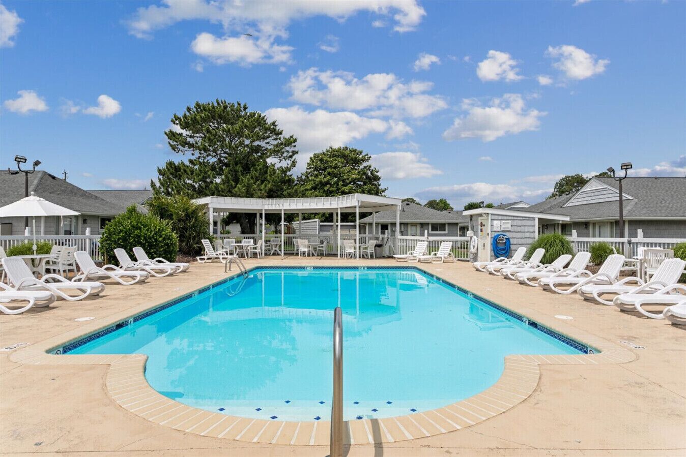 A rectangular swimming pool with a clear blue water, surrounded by white lounge chairs and a covered seating area.