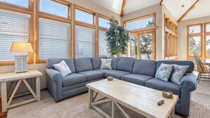 A living room with a sectional sofa, a wooden coffee table, and a lamp on a side table. Large windows provide natural light.