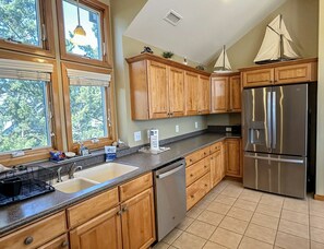 A kitchen with wooden cabinets, a stainless steel refrigerator, a dishwasher, and a window with a view of trees. - Full Catch: Well appointed with generous amenities! (Ocracoke)