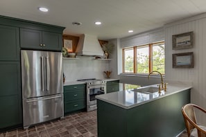 A kitchen with green cabinets, stainless steel appliances, a central island with a sink, and a window with a view.