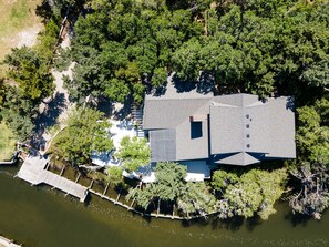 An aerial view of a house with a large deck overlooking a body of water, surrounded by dense trees.