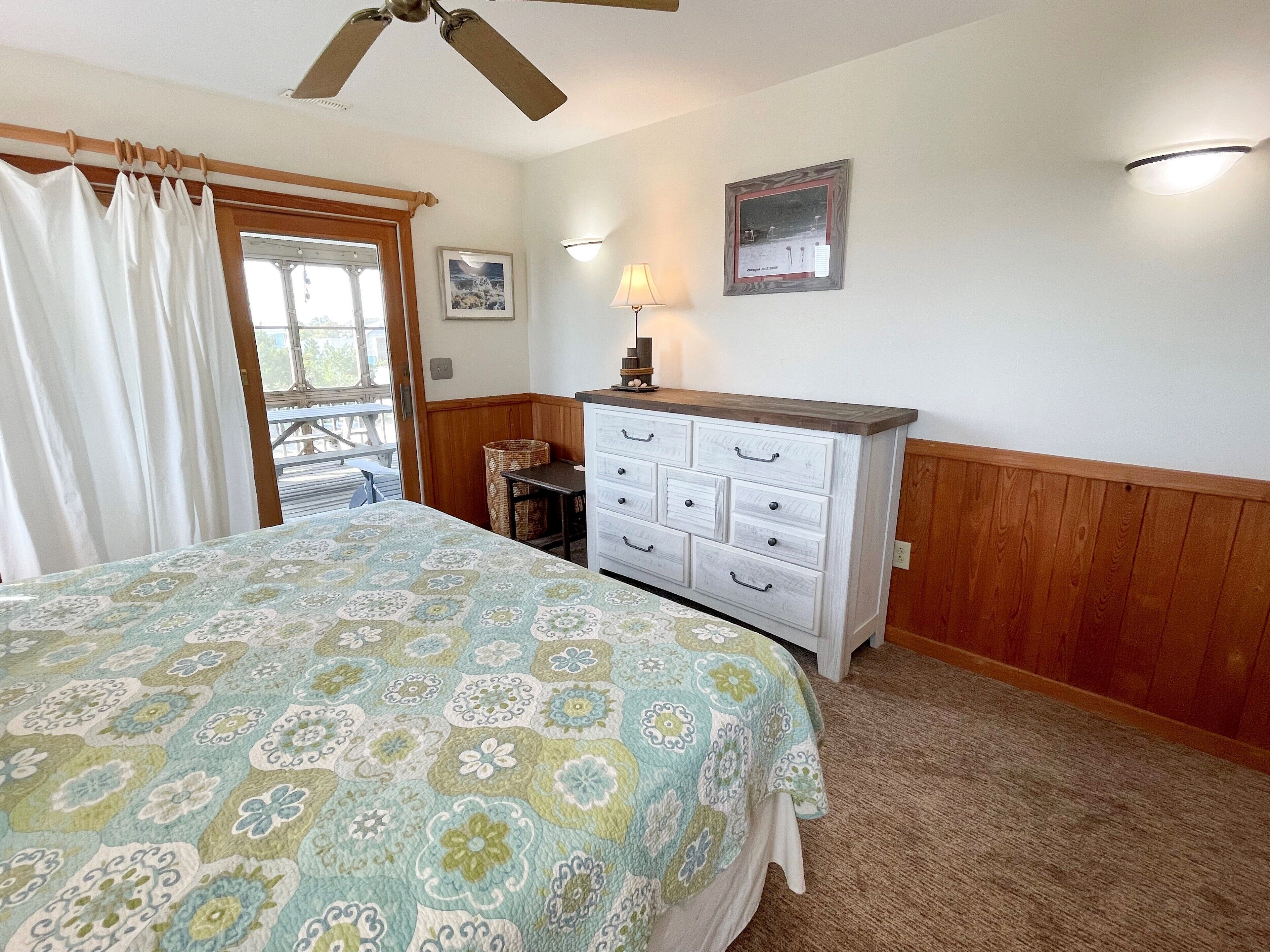 A bedroom with a bed, a dresser, a ceiling fan, a window with curtains, a wall-mounted lamp, and a framed picture.