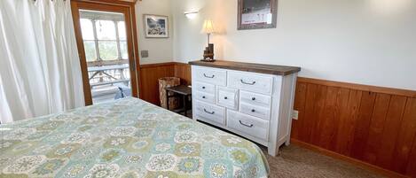 A bedroom with a bed, a dresser, a ceiling fan, a window with curtains, a wall-mounted lamp, and a framed picture.