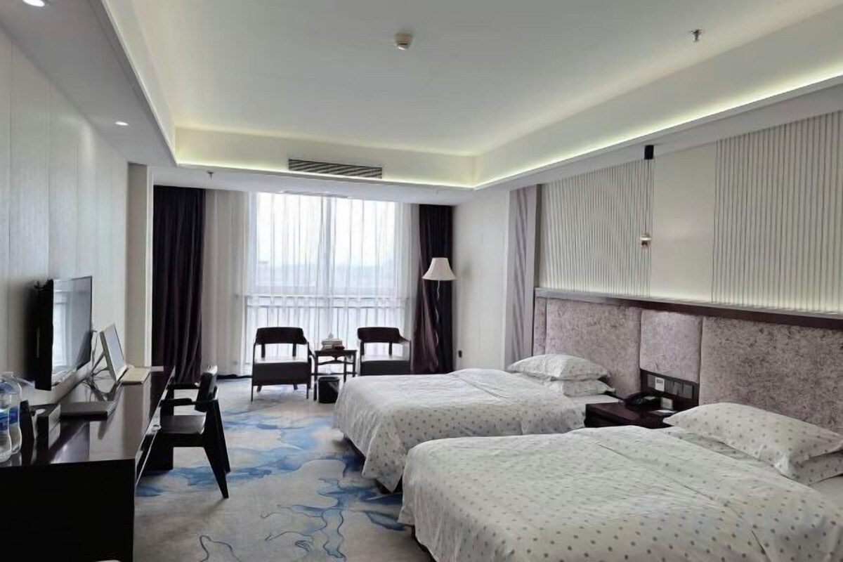 A hotel room with two beds, a desk, a chair, a TV, and a window with curtains.