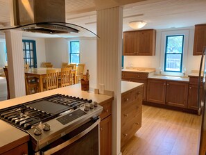 A modern kitchen with wooden cabinets, a stainless steel stove, and a dining area with wooden chairs. - Miss Elecia Garrish Home: Dog friendly, centrally located (Ocracoke)