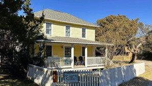 Une maison jaune à deux étages, dotée d’un porche et entourée d’une clôture blanche en piquets.