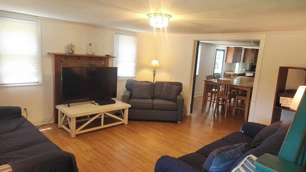 A living room with a TV, sofa, and a dining area with chairs and a table. - Miss Elecia Garrish Home: Dog friendly, centrally located (Ocracoke)