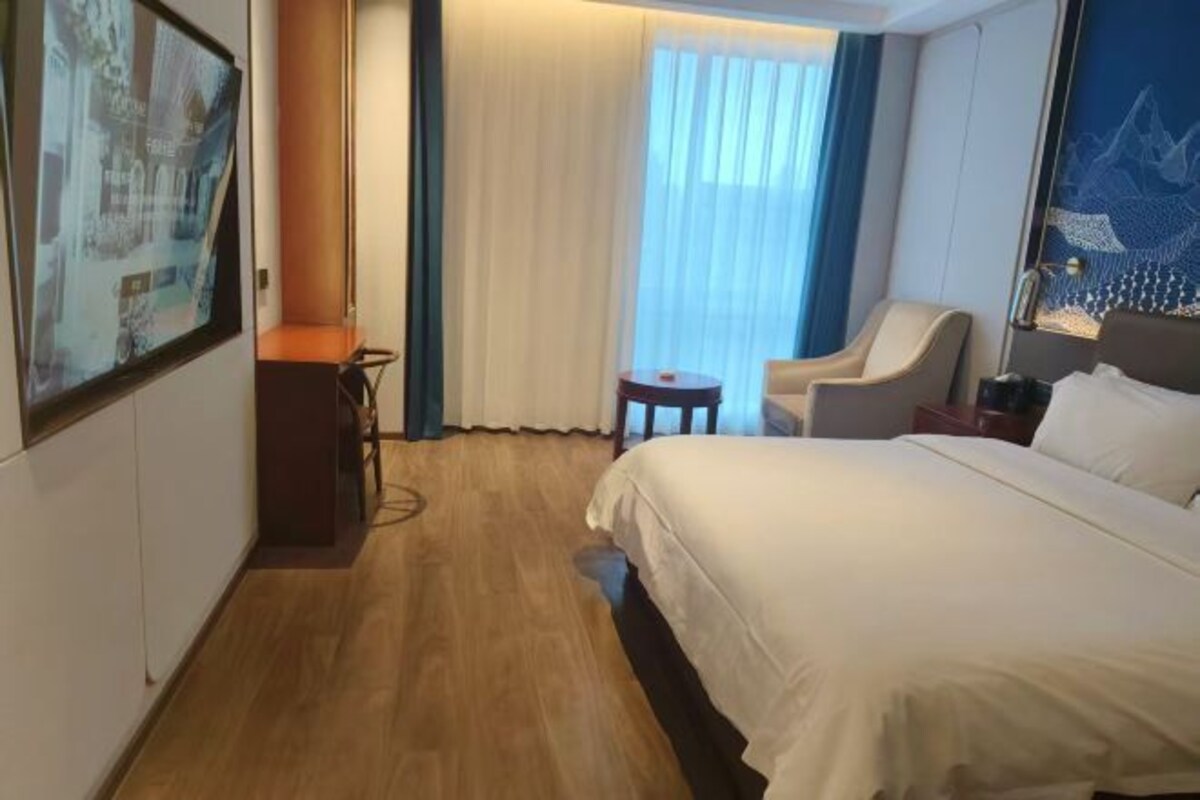 Una habitación de hotel con una cama grande, una silla, una mesita pequeña y un ventanal con cortinas.