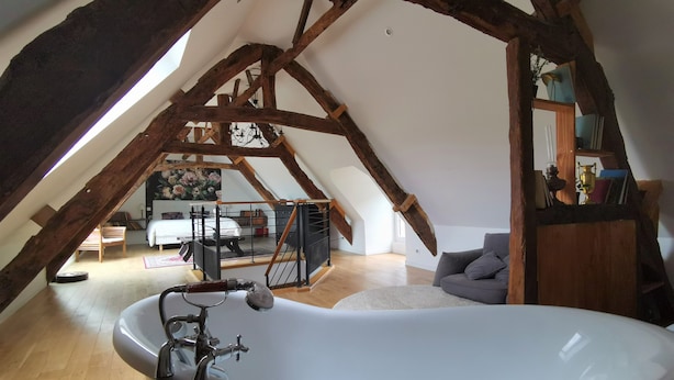 Een zolderkamer met een groot bad, houten balken, een bed en een boekenplank.