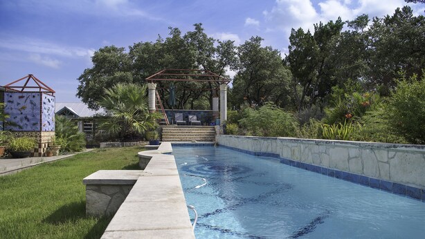 Ein Pool mit Steinrand, einer überdachten Sitzgelegenheit und üppiger Bepflanzung im Hintergrund.