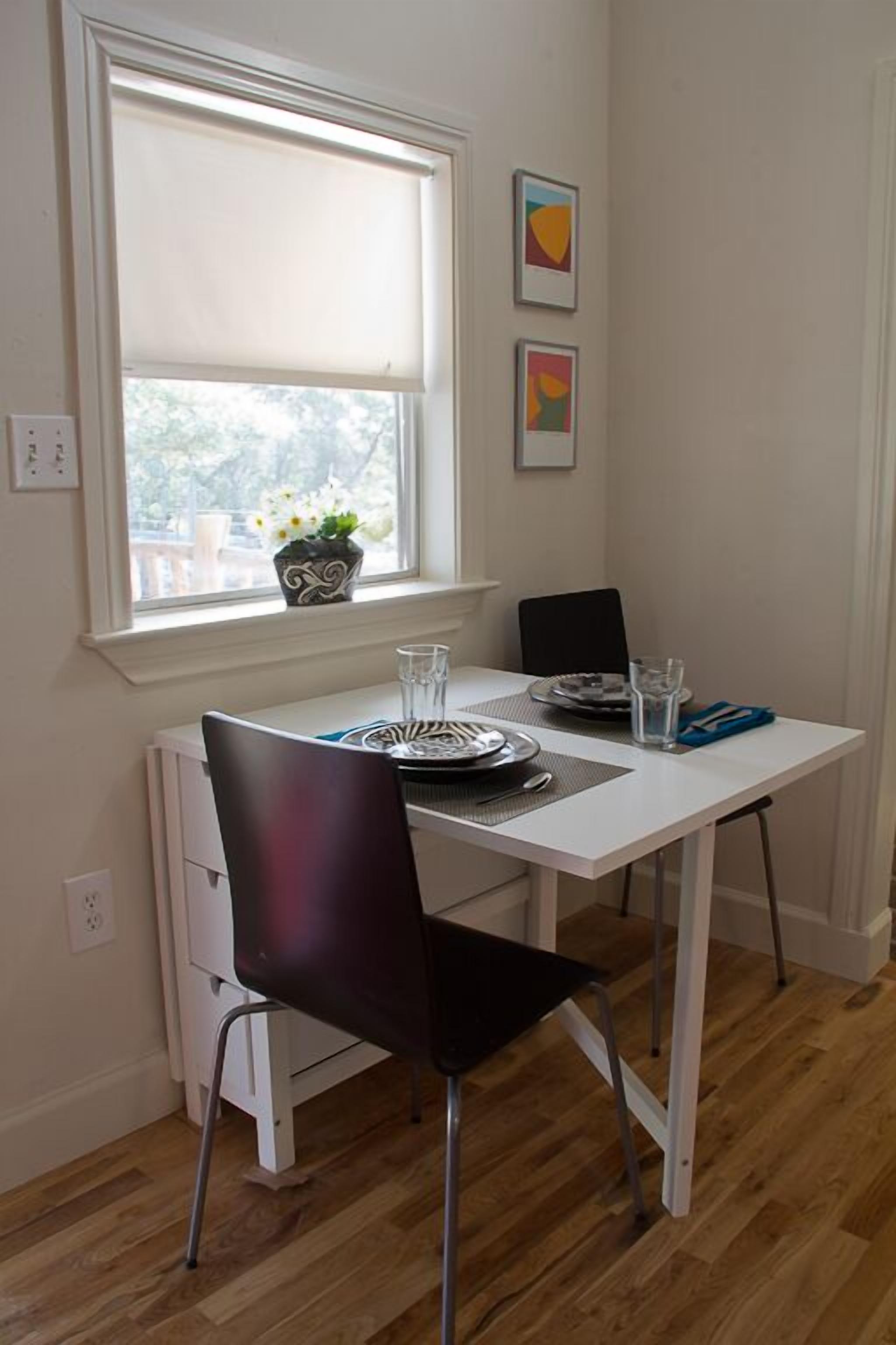 Un coin repas avec une table blanche, des chaises noires et une fenêtre donnant sur des arbres.