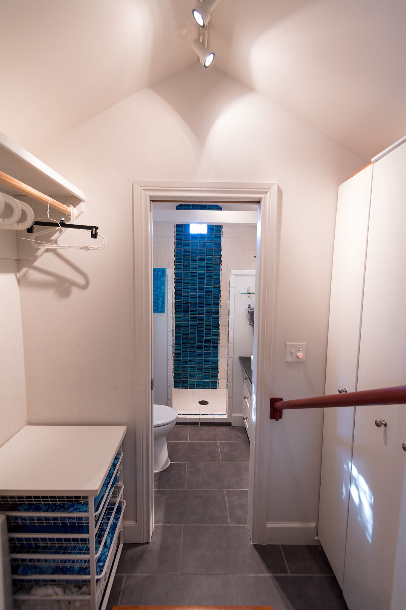 Un pasillo estrecho con una puerta blanca que da a un baño con azulejos azules. Hay armarios blancos y un banco de madera.