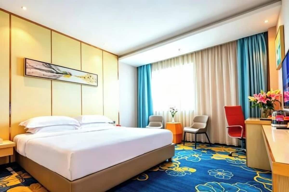 Habitación de hotel con cama, escritorio, dos sillas, un cuadro en la pared y ventana con cortinas.