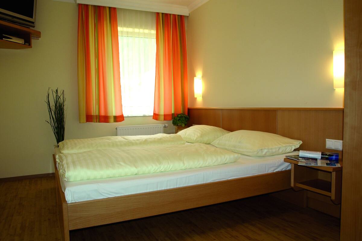 Una habitación de hotel con una cama de madera, una mesita de noche, una lámpara y un ventanal con cortinas.