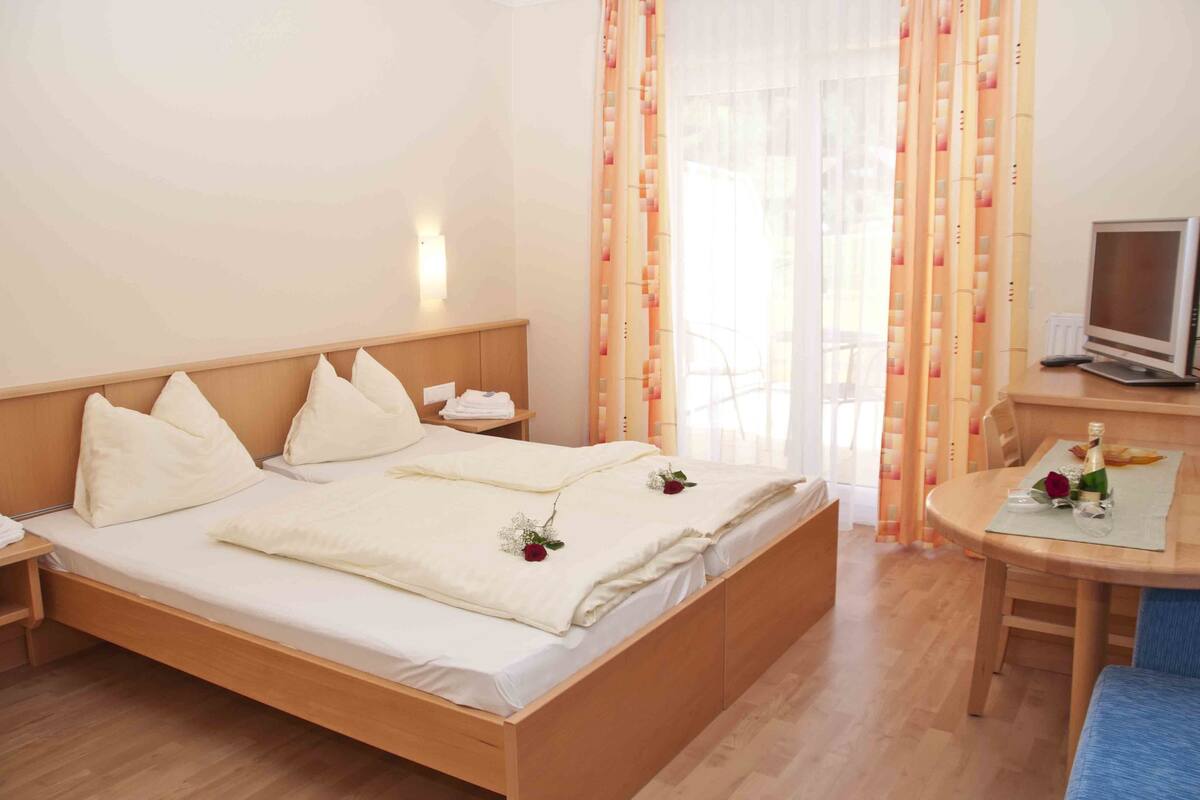 Habitación de hotel con cama de madera, mesita de noche con lámpara, un escritorio pequeño con una computadora y una ventana con cortinas.
