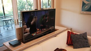 Un soggiorno con una grande televisione a schermo piatto montata su un supporto in legno, un divano bianco con cuscini decorativi e un tavolino con un portatile.