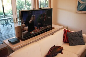 Una sala de estar con una televisión de pantalla plana grande montada sobre un soporte de madera, un sofá blanco con almohadones decorativos y una mesita con una computadora portátil.
