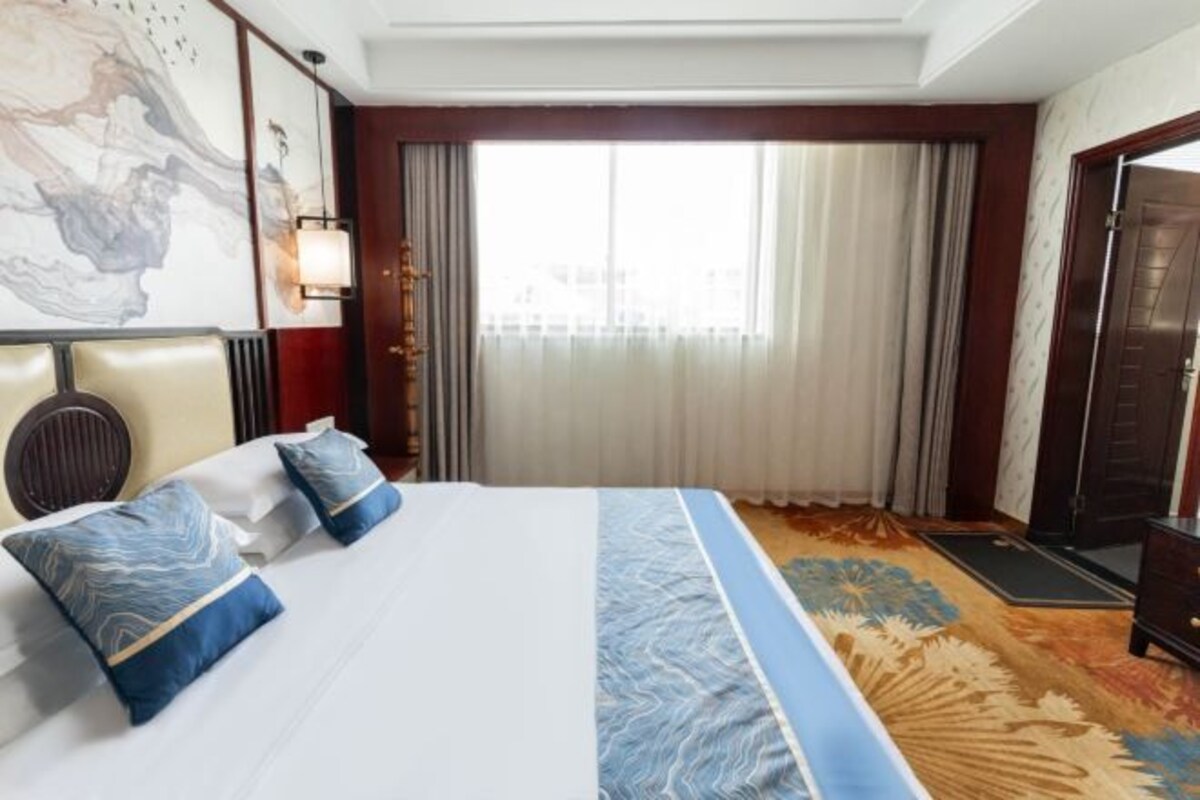 Une chambre d’hôtel avec un grand lit, deux oreillers, une tête de lit, une fenêtre avec des rideaux, une lampe fixée au mur et une moquette à motifs.