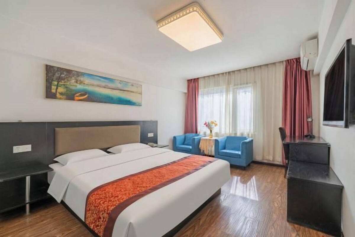 A hotel room with a bed, a desk, a chair, a sofa, a painting on the wall, and a window with curtains.