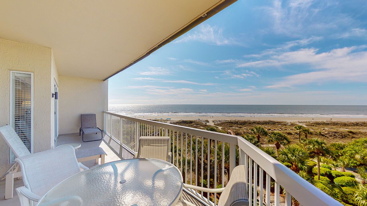 A balcony with a glass table and chairs, overlooking a beach with palm trees and a clear sky.