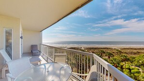 Ein Balkon mit Glastisch und Stühlen, der auf einen Strand mit Palmen und wolkenlosen Himmel blickt.