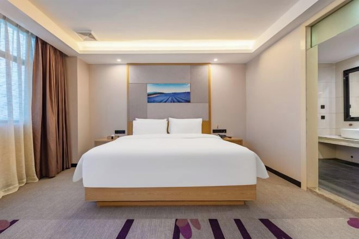 A modern hotel room with a large bed, a nightstand, a painting on the wall, and a bathroom visible through an open door.
