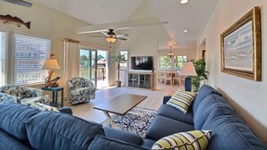A spacious living room with a blue sofa, a coffee table, and a TV. There is a ceiling fan, a chandelier, and a large window with blinds.