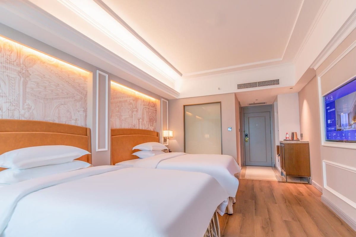 A hotel room with two beds, a wooden floor, a wall-mounted TV, and a door leading to a hallway.