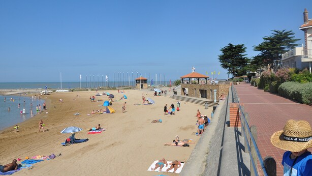 Une plage où des gens se prélassent au soleil et se baignent, une tour de surveillance des sauveteurs et un bâtiment avec un toit rouge.