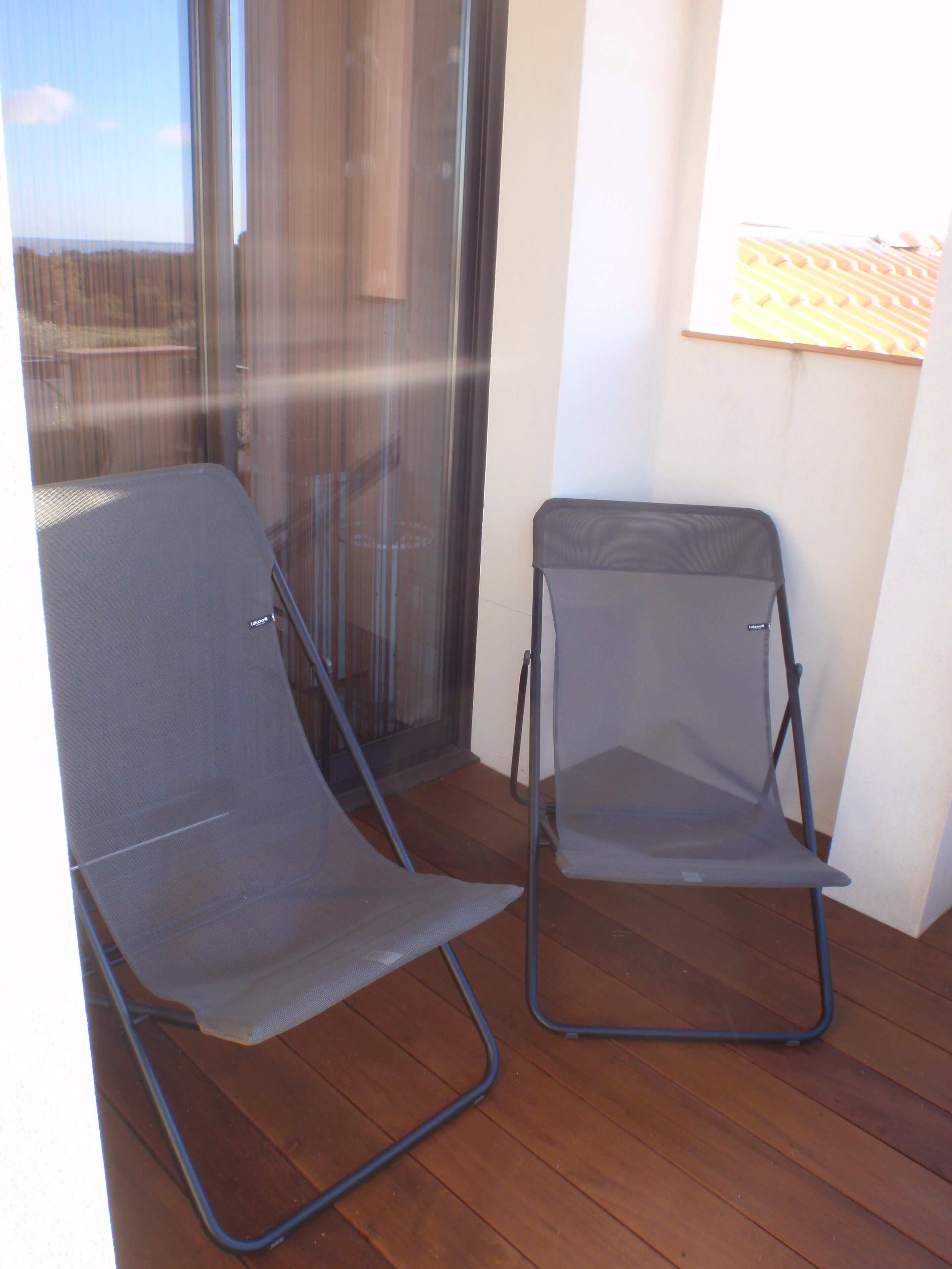 Deux chaises pliantes sur une terrasse en bois, avec une porte vitrée et un mur blanc orné d’un tableau encadré.