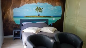 Une chambre à coucher comprenant un lit, deux chaises, une table de chevet et une fresque murale représentant une scène de plage.