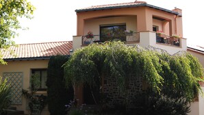 Une maison à deux étages, avec un toit recouvert de tuiles rouges, un mur en pierre et un jardin luxuriant où poussent diverses plantes et fleurs.