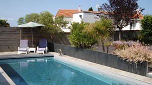 Une piscine rectangulaire, agrémentée de deux chaises longues et d’un parasol, entourée d’un petit muret en pierre et d’une végétation luxuriante.