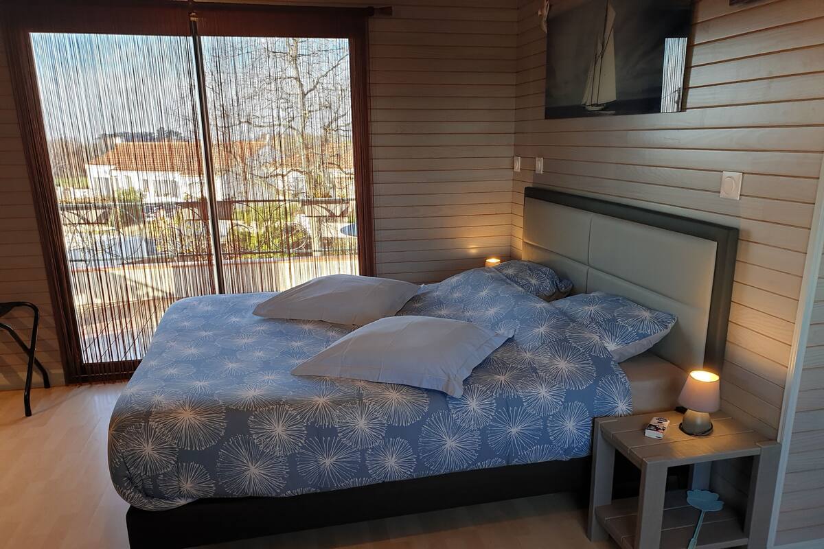 Une chambre à coucher avec un lit, des oreillers, une table de chevet avec une lampe et une fenêtre avec des stores.
