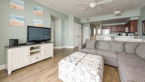 A living room with a TV, sofa, ottoman, and artwork on the wall. - Elite Beach Retreat - Ocean Walk (Daytona Beach)