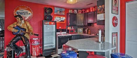 A retro-themed kitchen with a checkered floor, red and black cabinets, a Coca-Cola refrigerator, and a jukebox statue.