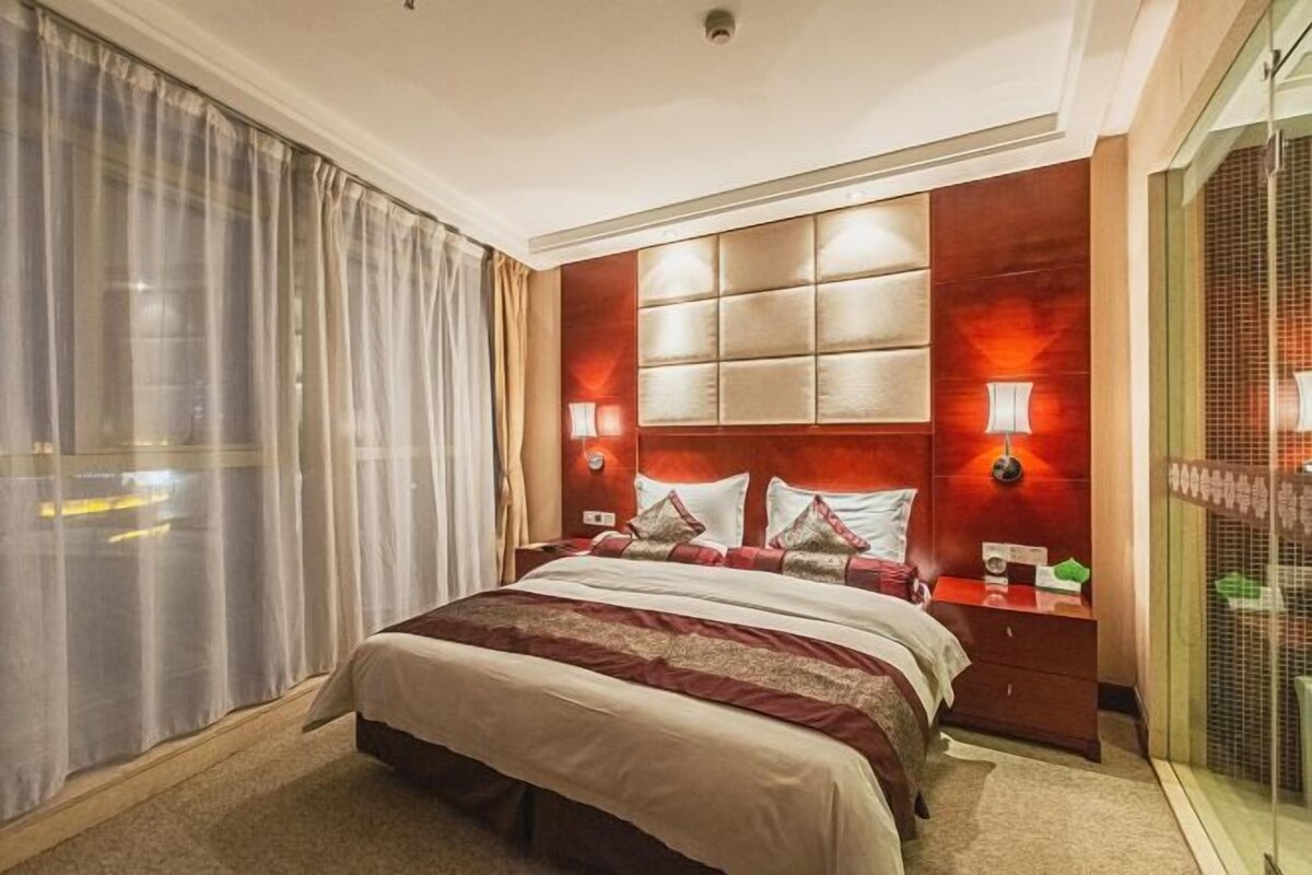 Une chambre d’hôtel avec un grand lit, une tête de lit dotée de panneaux lumineux, une table de chevet avec une lampe et une fenêtre ornée de rideaux légers.