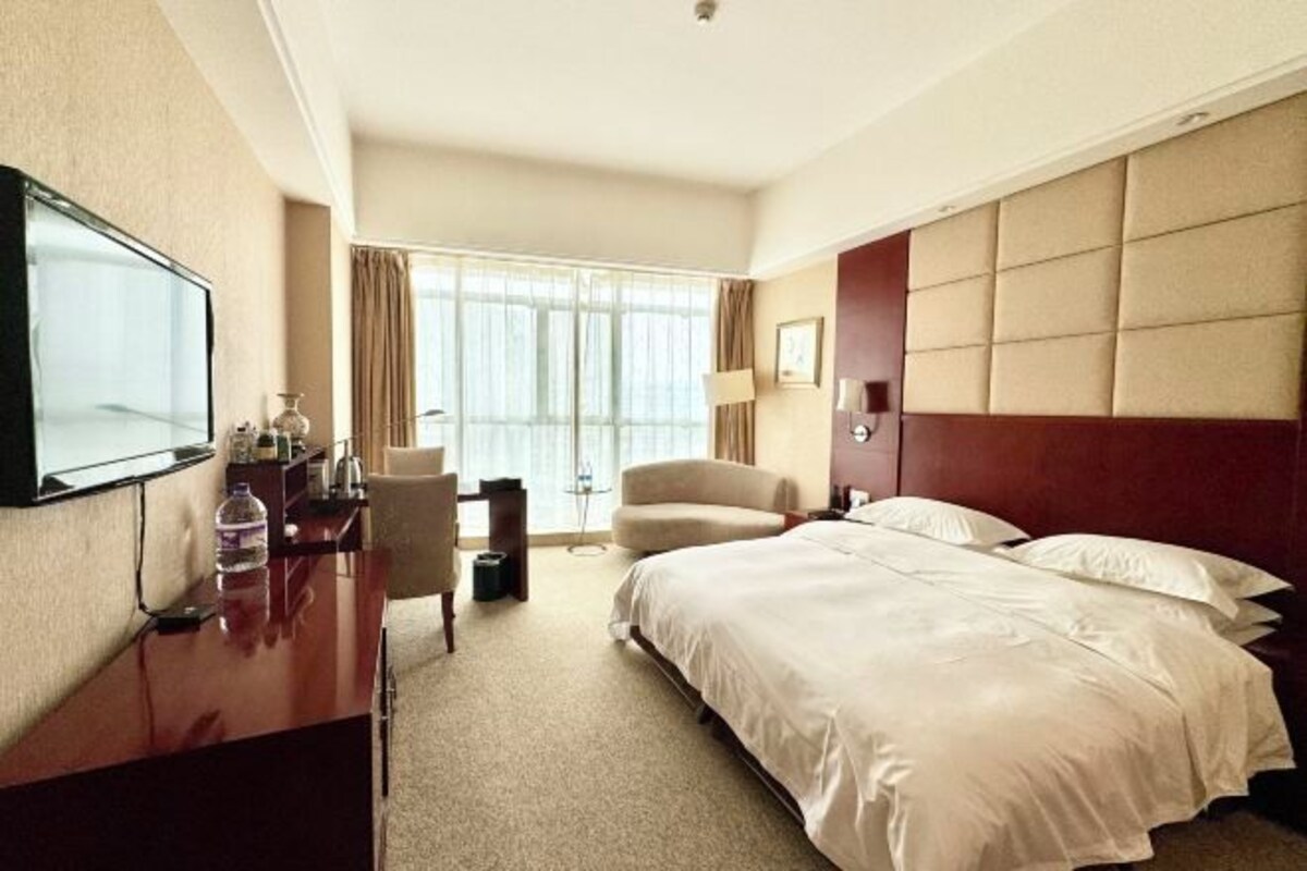 Een hotelkamer met een bed, een bureau met stoel, een televisie en een groot raam met gordijnen.
