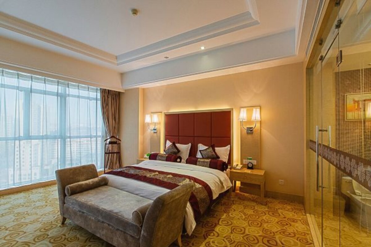 Een hotelkamer met een groot bed, een bank, een nachtkastje met een lamp, een douchecabine van glas en een groot raam met gordijnen.