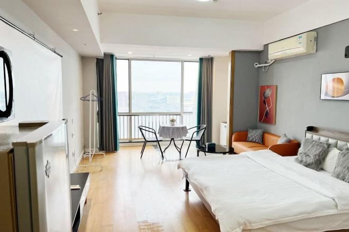 A modern hotel room with a large bed, a dining area with a table and chairs, a balcony with a view, and a wardrobe.