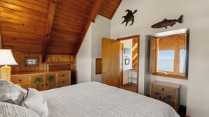 Ein Schlafzimmer mit Holzbalkendecke, einem Bett mit weißer Bettwäsche, einem hölzernen Schminktisch und einem Spiegel, mit Blick auf das Meer durch ein Fenster.