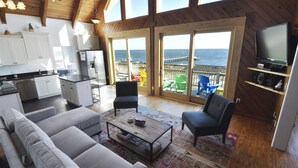Ein großzügiger Wohnbereich mit einer großen Couch, einem Couchtisch und einem Fernseher. Es gibt eine Küchenzeile mit Schränken und einem Kühlschrank. Das Zimmer verfügt über große Fenster mit Blick auf den Ozean.