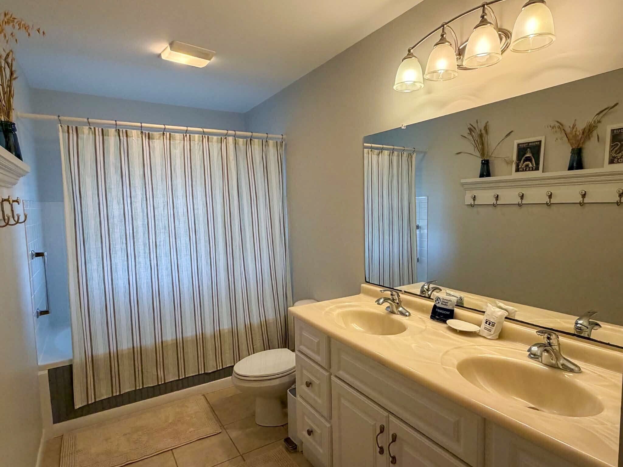 Une salle de bain avec un rideau de douche, deux lavabos, des toilettes et un miroir avec une étagère au-dessus.