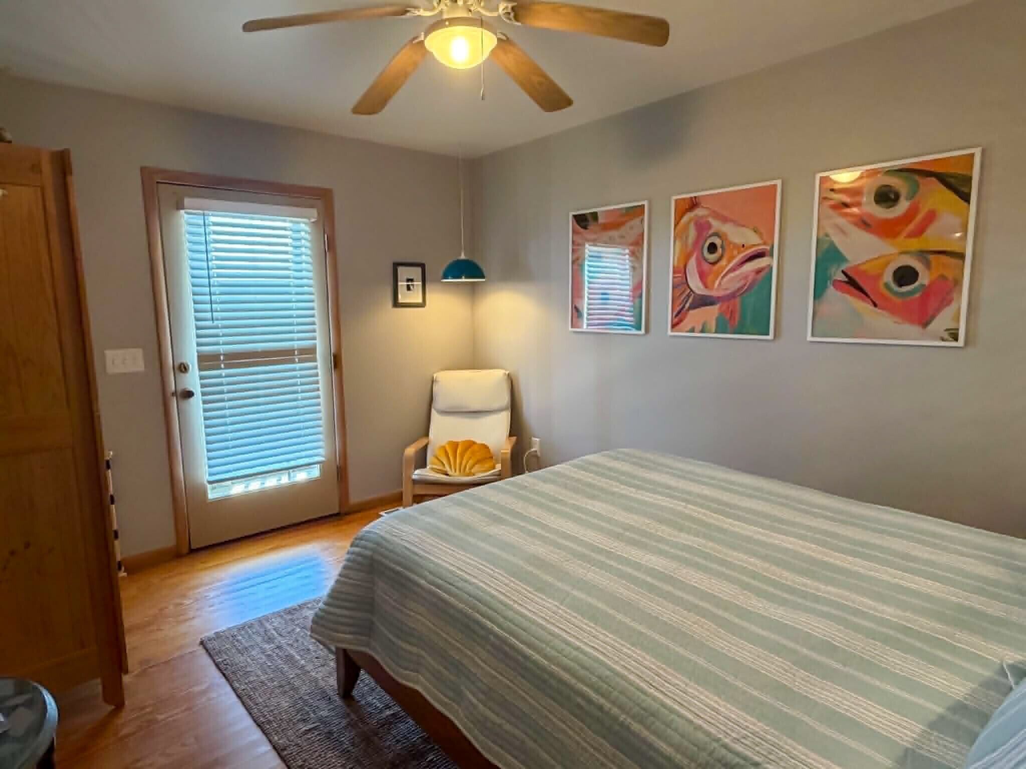 A bedroom with a bed, a chair, a ceiling fan, and three wall-mounted paintings.
